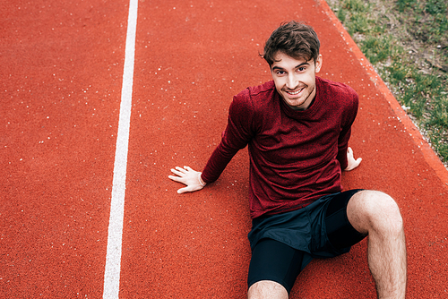Handsome sportsman smiling at camera while sitting on running track outdoors