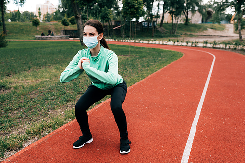 Sportswoman in medical mask doing squat on running path in park