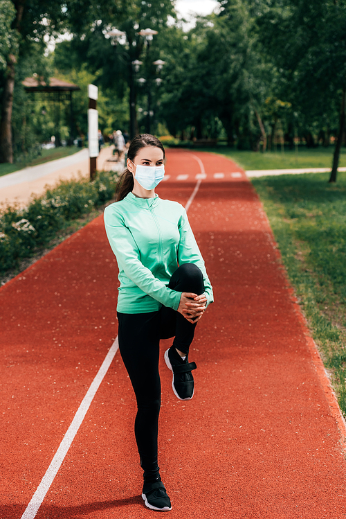Sportswoman in medical mask stretching leg on running track in park