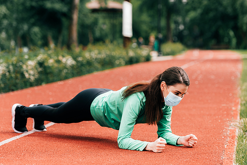 Side view of sportswoman in medical mask doing plank while working out in park