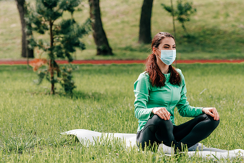 Woman in medical mask sitting on fitness mat in park