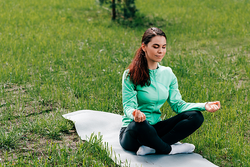 Beautiful woman with crossed legs meditating on fitness mat on grass in park