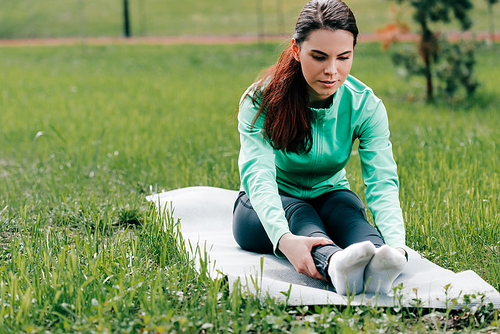 Selective focus of attractive sportswoman stretching on fitness mat on grass in park