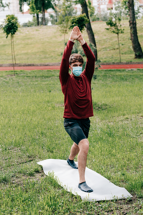 Sportsman in medical mask with raised hands training on fitness mat in park