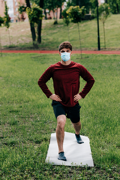Young sportsman in medical mask with hands on hips exercising on fitness mat in park