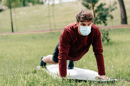 Sportsman in medical mask doing push ups during working out on fitness mat on grass in park