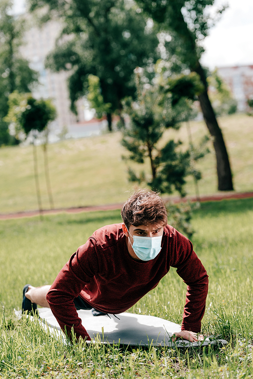 Selective focus of young sportsman in medical mask doing press ups on fitness mat on grass in park