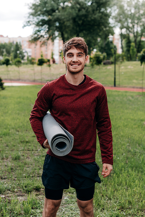 Handsome sportsman holding fitness mat and smiling at camera in park