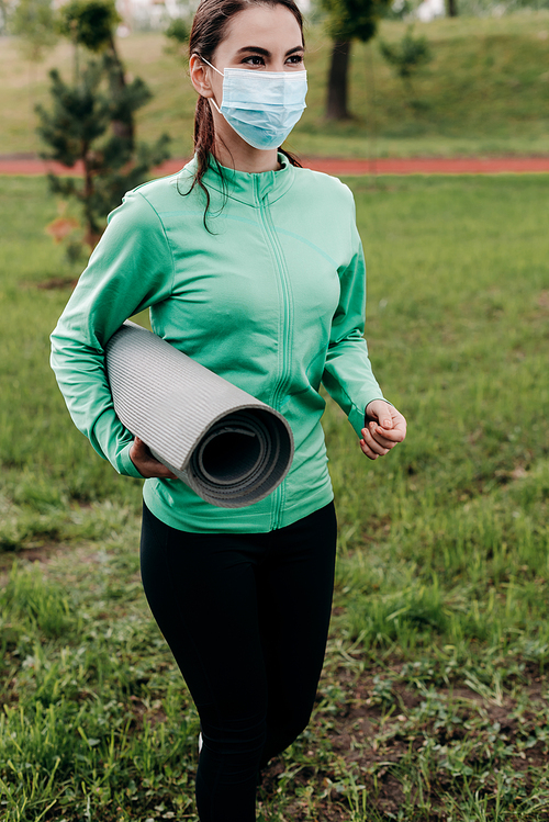 Young sportswoman in medical mask holding fitness mat while walking in park