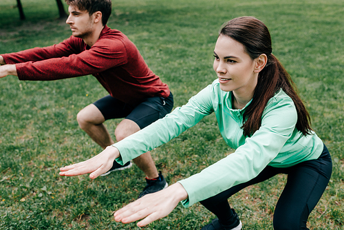 Selective focus of smiling sportswoman doing squat while working out near boyfriend in park