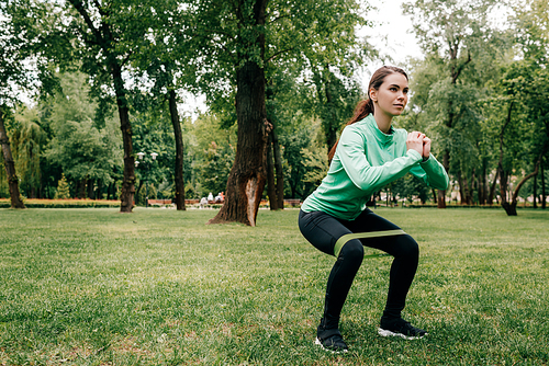 Attractive sportswoman doing squat with resistance band in park