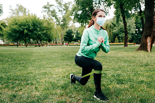 Young sportswoman in medical mask training with resistance band in park
