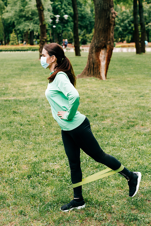 Side view of sportswoman in medical mask using resistance band during training in park