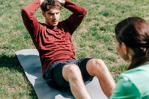 Selective focus of handsome man doing abs near girlfriend on fitness mat in park