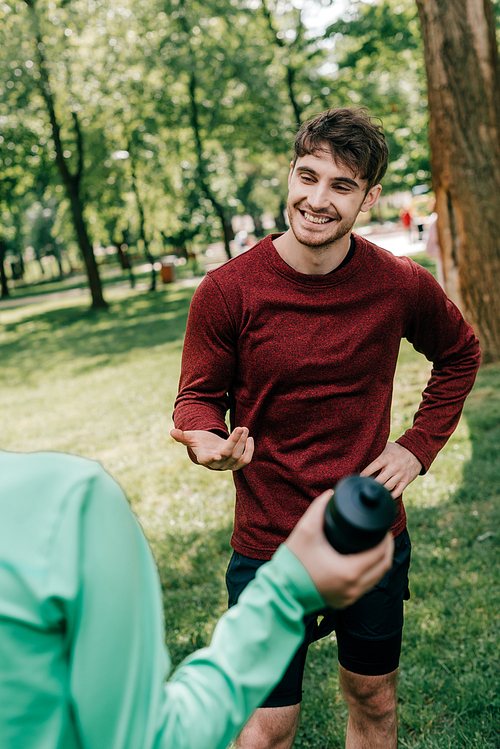 Selective focus of handsome sportsman talking to girlfriend with sports bottle in park