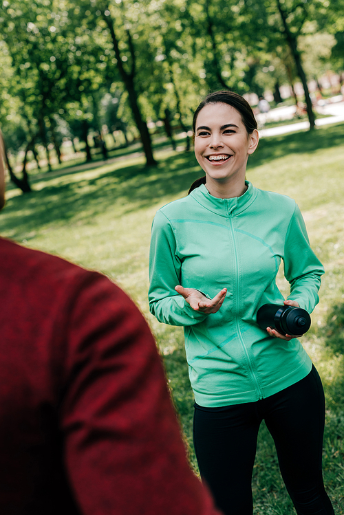 Selective focus of smiling sportswoman talking to boyfriend while holding sports bottle in park