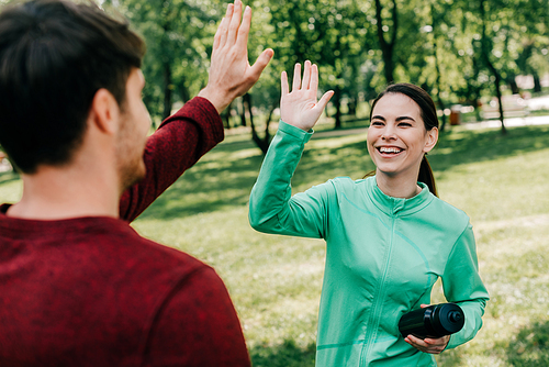 Selective focus of smiling sportswoman with sports bottle high five to boyfriend in park