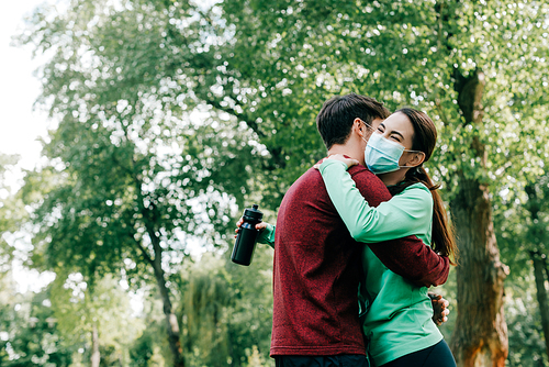 sportsman embracing girlfriend in medical mask holding sports bottle in park