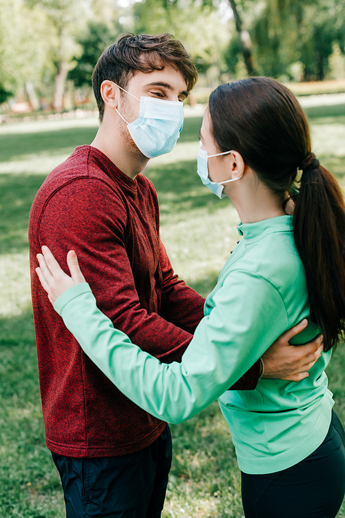 Side view of sportswoman in medical mask embracing boyfriend in park
