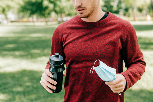 Cropped view of sportsman holding sports bottle and medical mask in park