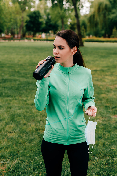 Beautiful sportswoman drinking water and holding medical mask in park