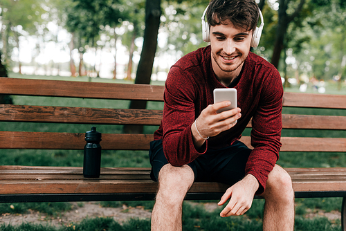 Smiling sportsman in headphones using smartphone near sports bottle on bench in park