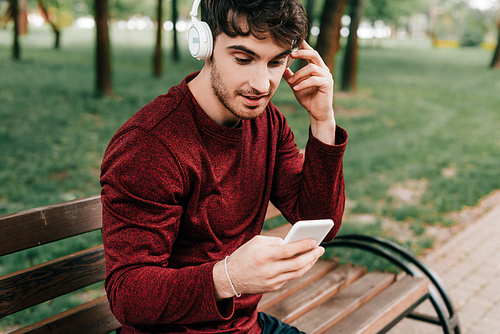 Handsome sportsman listening music in headphones and using smartphone on bench in park