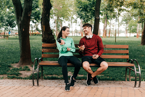 Smiling sportsman in headphones looking at girlfriend with sports bottle on bench in park