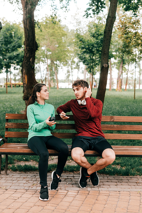 Smiling sportswoman holding sports bottle near boyfriend in headphones with smartphone on bench in park