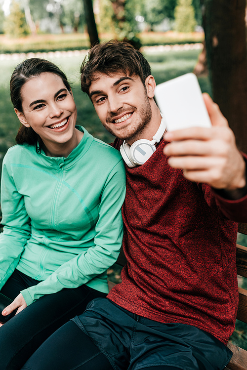 Selective focus of smiling sportsman taking selfie with smartphone near girlfriend on bench in park