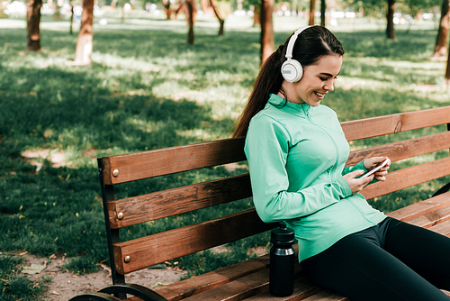 Smiling sportswoman in headphones using smartphone near sports bottle on bench in park