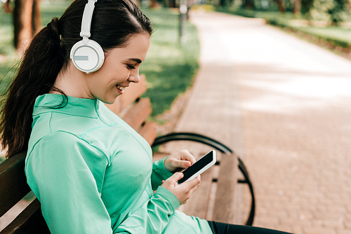 Side view of smiling sportswoman in headphones using smartphone on bench in park