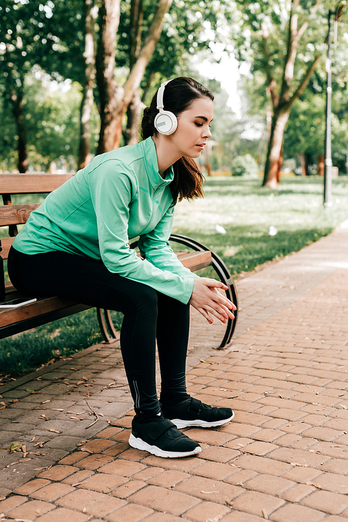 Side view of young sportswoman in headphones resting on bench in park