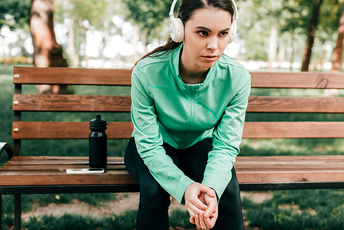 Sportswoman in headphones sitting near smartphone and sports bottle on bench in park