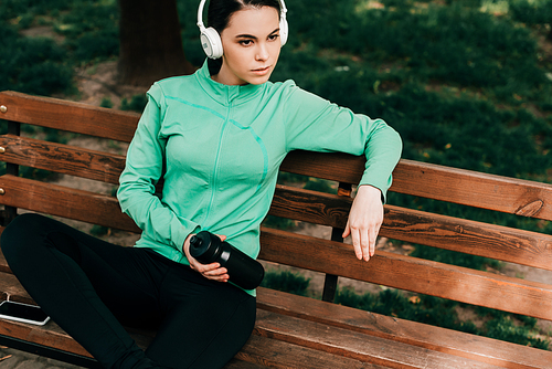 Beautiful sportswoman in headphones holding sports bottle while resting on bench in park