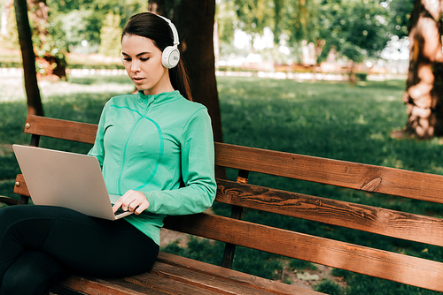Woman in headphones using laptop while sitting on bench in park