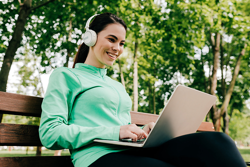 Smiling girl in headphones using laptop on bench in park