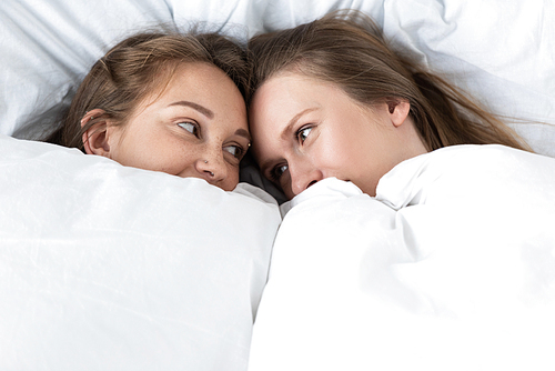 two lesbians lying under white blanket and looking at each other in bedroom