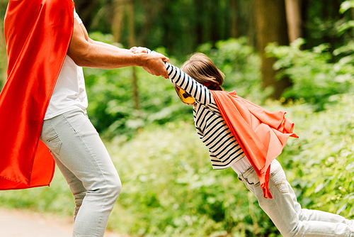 cropped view of father spinning around son in forest while boy holding hands of dad