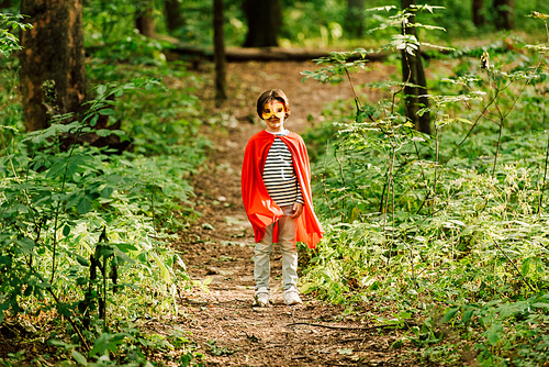 full length view of small boy standing in superhero costume in forest on path