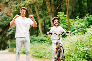 father smiling and cheering son while boy riding bicycle and 