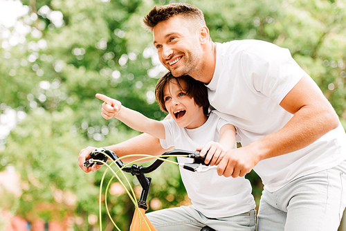 excited father and son looking forward while boy pointing with finger and dad helping kid to ride on bicycle