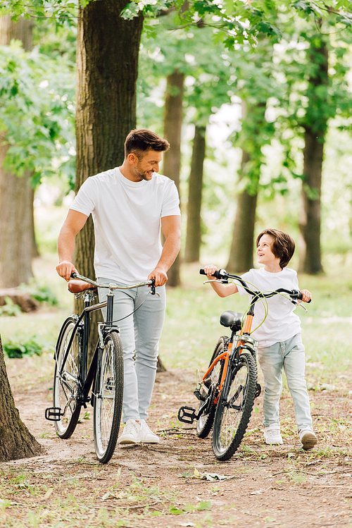 full length view of happy father and son looking at each other and smiling while walking with bicycles in forest