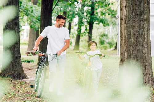 selective focus of father and son smiling while walking in park with bicycles