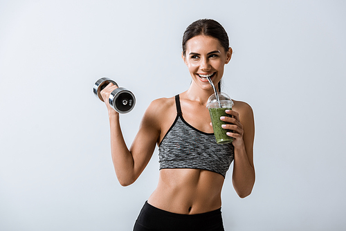 smiling sportswoman holding dumbbell and drinking smoothie isolated on grey
