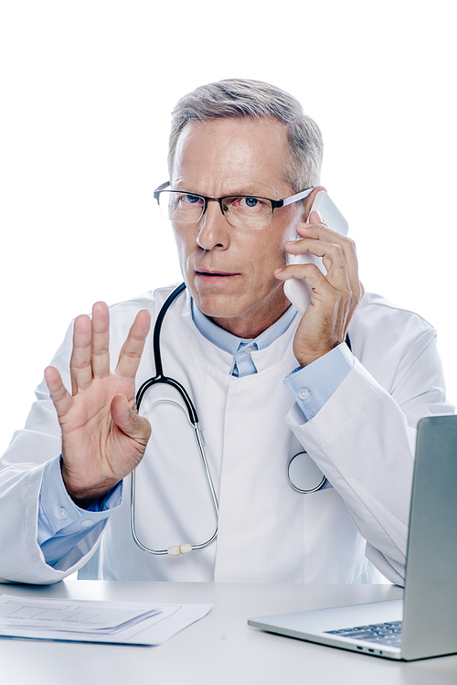 handsome doctor in white coat talking on smartphone and showing gesture isolated on white