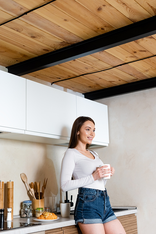 happy girl holding cup of tea near plate with croissant on table