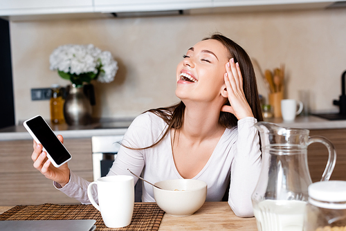happy woman holding smartphone with blank screen and laughing near breakfast