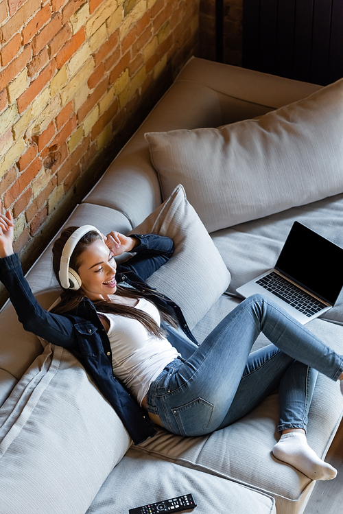 overhead view of happy woman listening music in wireless headphones near laptop with blank screen