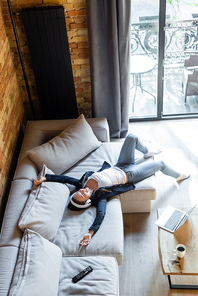 overhead view of happy woman in wireless headphones listening music near laptop and remote controller while lying on sofa